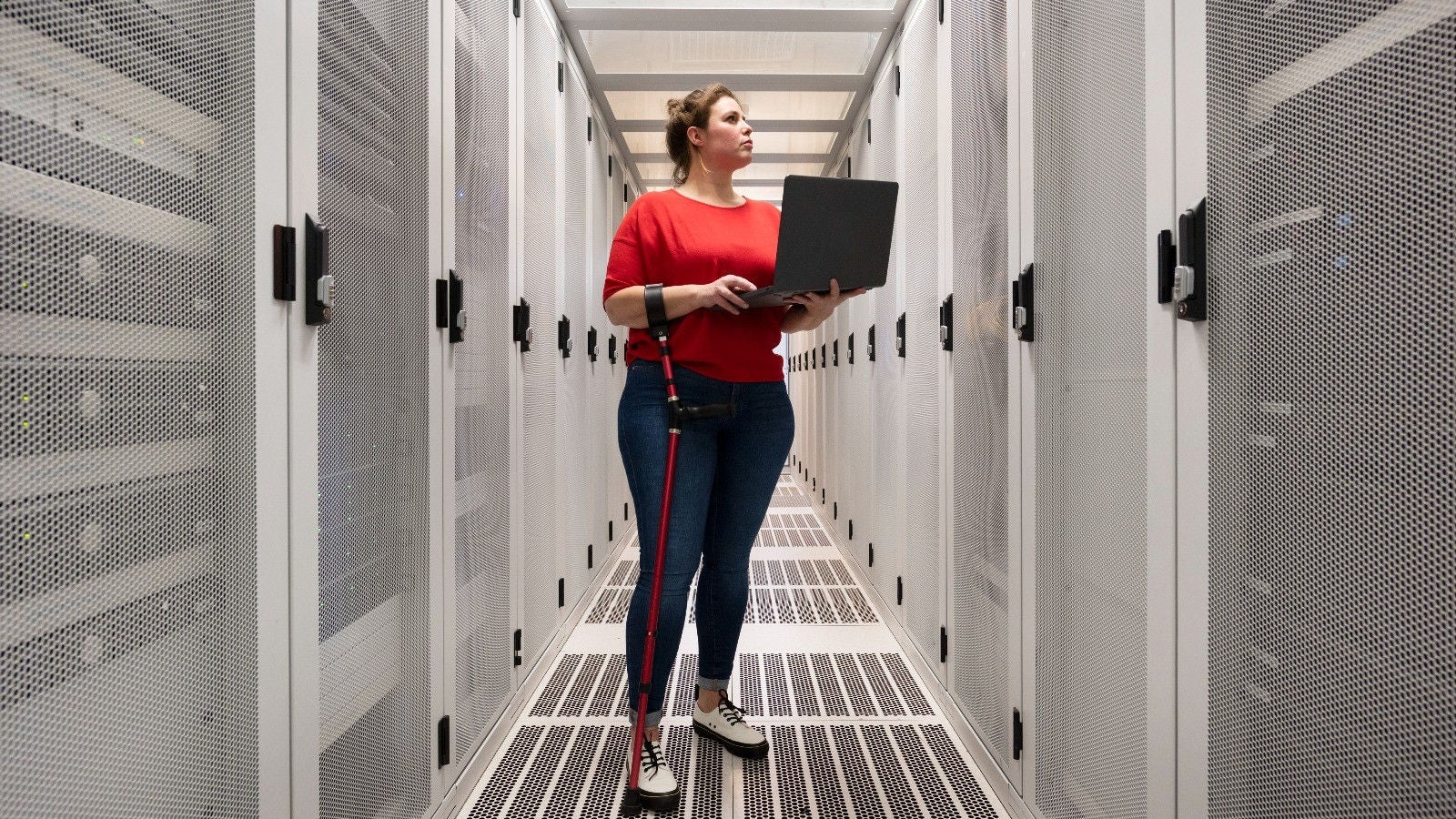 Woman with a walking cane holding a laptop while inspecting servers in a data centre aisle.