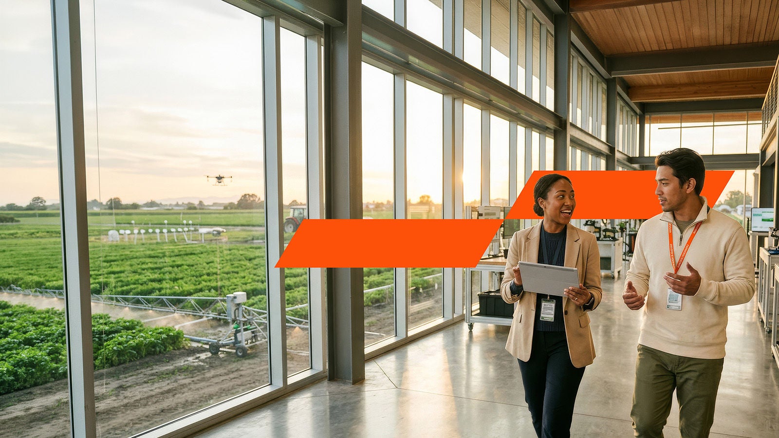 Two professionals walking in glass corridor with tablet; farmland and drone outside.